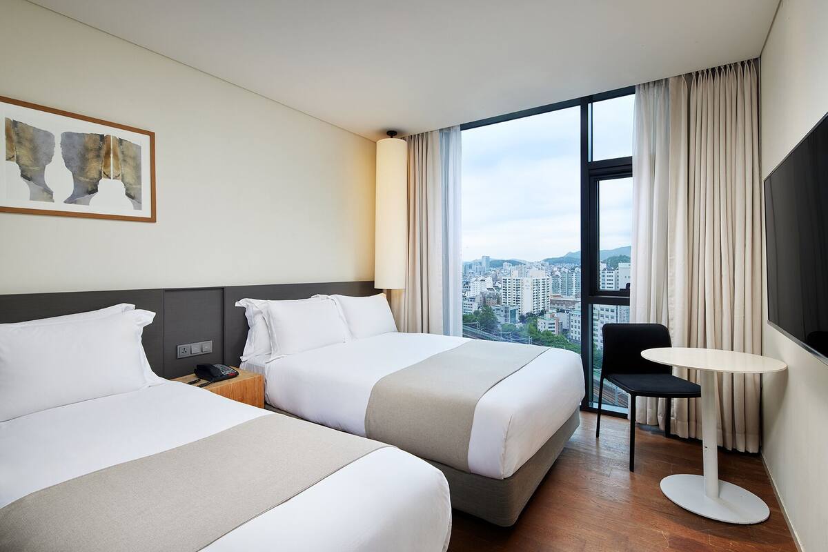 A hotel room with two beds, a desk, a chair, a TV, and a window with a city view.