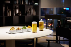 Una mesa redonda cubierta con un mantel blanco, tres tazones pequeños de comida y dos vasos de cerveza.