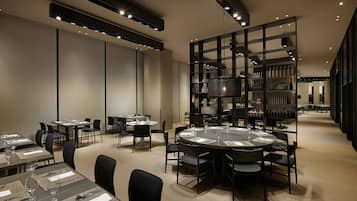 A modern dining area with round tables, black chairs, and a central chandelier.