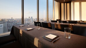 Un comedor con mesas dispuestas para dos, ventanas amplias con vista a la ciudad y una decoración de estilo moderno.