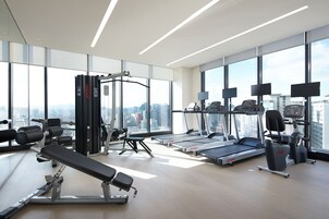 A modern gym with cardio machines, weight benches, and large windows offering a city view.