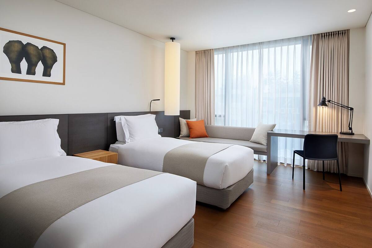 A hotel room with two beds, a desk, a chair, and a large window with curtains.