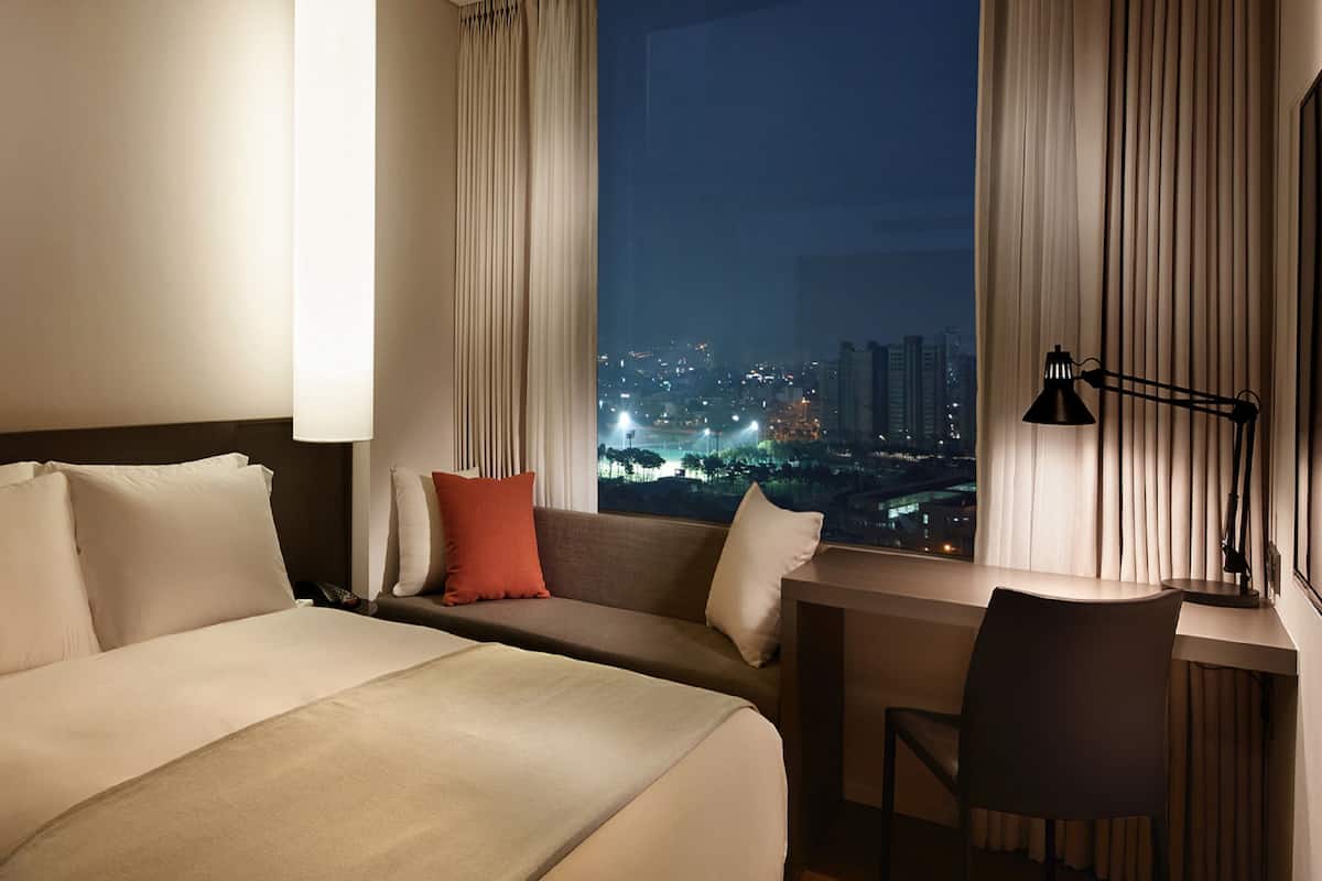 A hotel room with a bed, a desk, a chair, a window with curtains, and a city view at night.