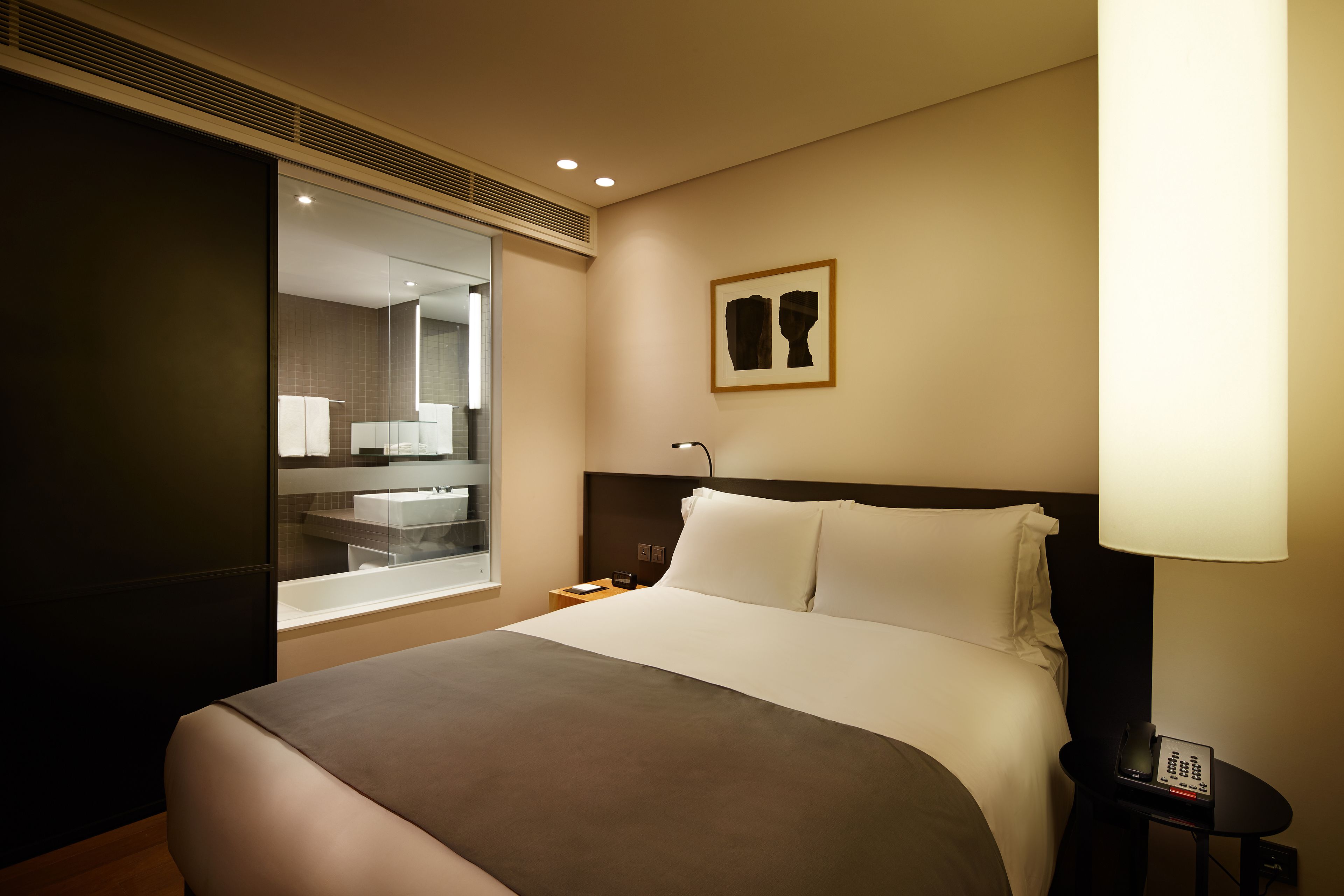 A hotel room with a large bed, a bedside table with a lamp, a mirror, and a framed artwork on the wall.