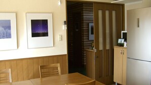 Een eethoek met een houten tafel en stoelen, een keuken met een koelkast en een magnetron, en twee ingelijste foto's aan de muur.