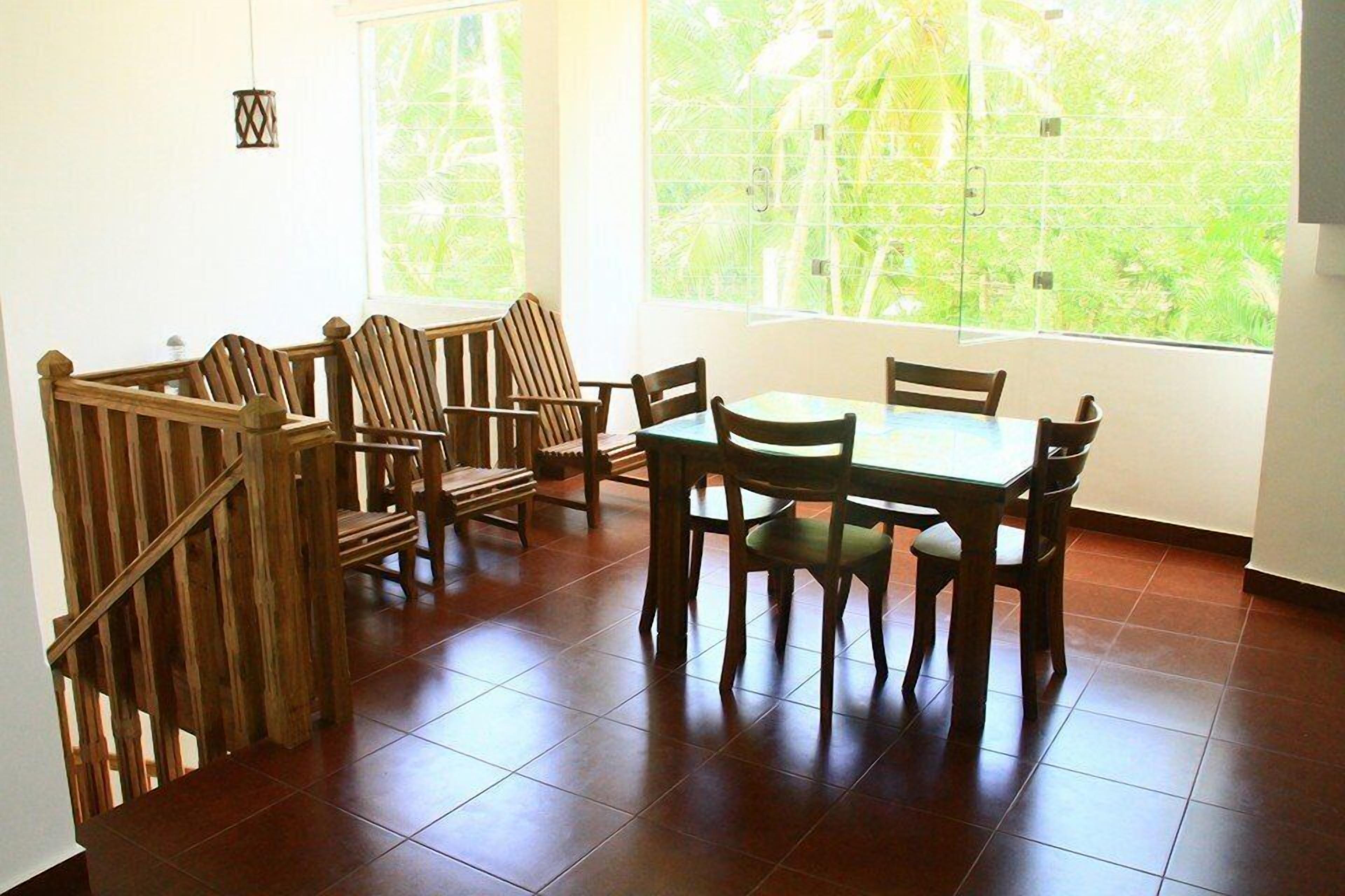 A dining area with a wooden table and chairs, large windows with a view of greenery, and a tiled floor.