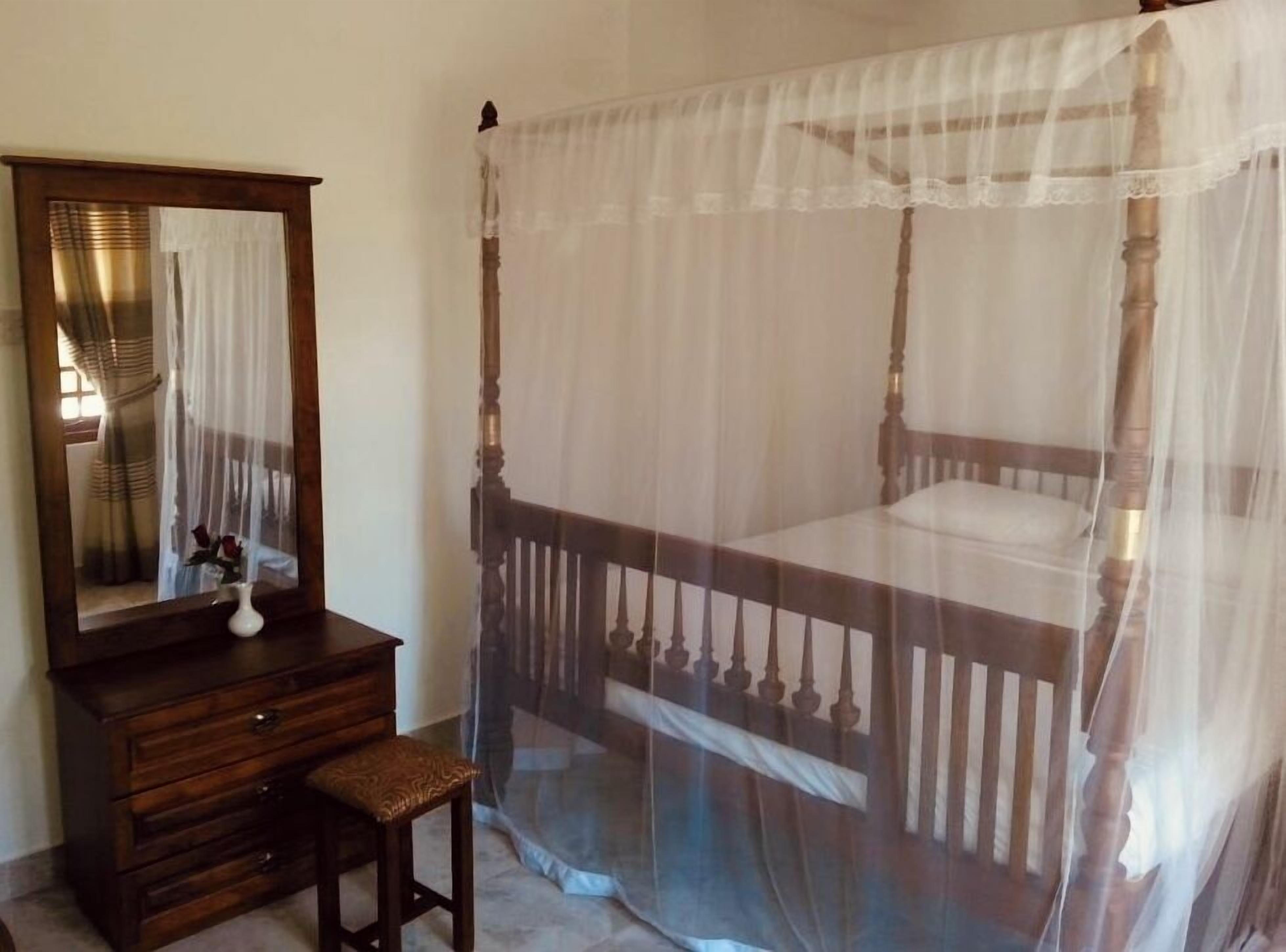 A bedroom with a four-poster canopy bed, a wooden dresser with a mirror, and a small chair.