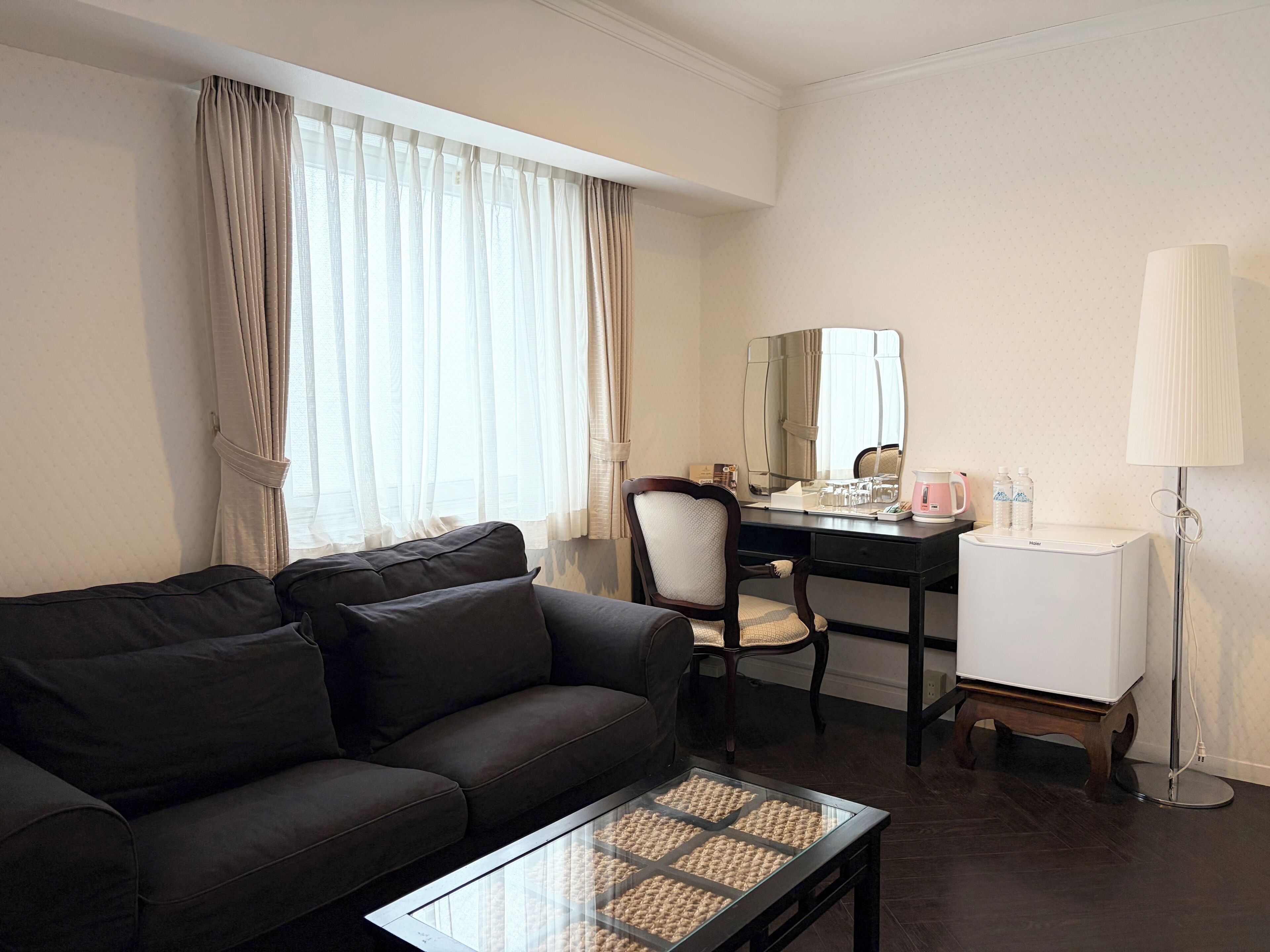 A hotel room with a dark brown sofa, a glass coffee table, a desk with a chair, a mirror, a lamp, and a minibar.