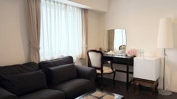 A hotel room with a dark brown sofa, a glass coffee table, a desk with a chair, a mirror, a lamp, and a minibar.