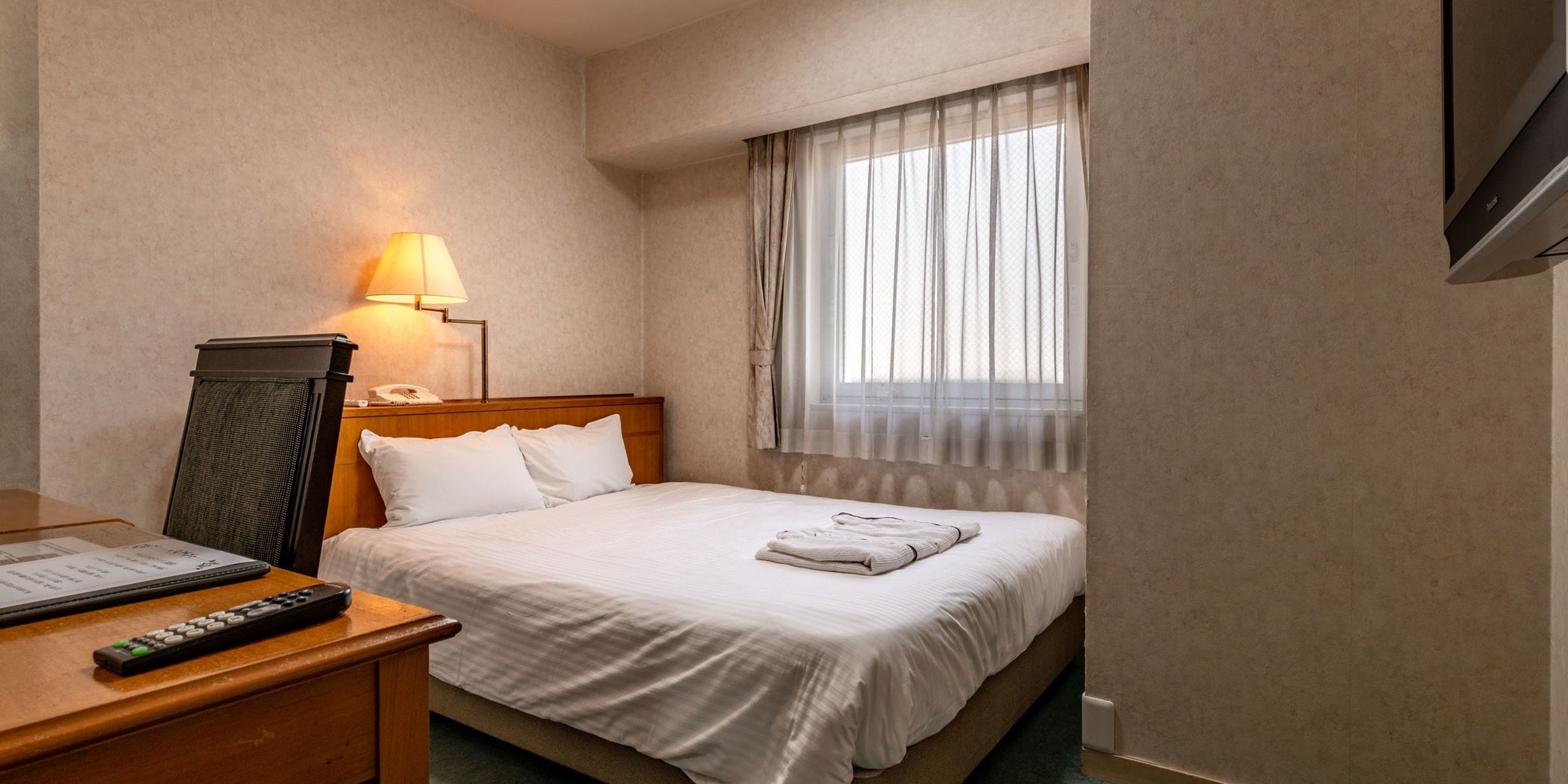 A hotel room with a bed, a desk with a lamp, a remote control, and a window with sheer curtains.