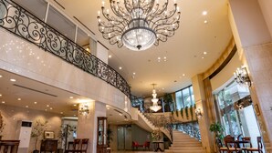 Um hall imponente com piso de mármore, uma escadaria monumental com grades elaboradas e um grande candelabro.