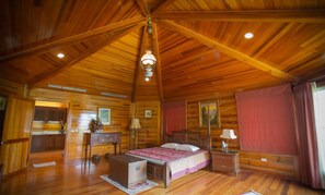 A log cabin bedroom with a wooden ceiling, a bed, a dresser, a desk, and a window with curtains.