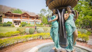A statue of two figures in green attire, a fountain, and a building with a wooden facade in the background. - The Log Home Experience Khao Yai (Pak Chong)