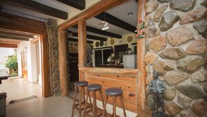 A rustic kitchen with a stone wall, wooden beams, and a bar area with stools. - The Log Home Experience Khao Yai (Pak Chong)