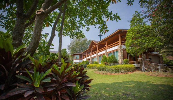 A large, log cabin-style house with a stone chimney, surrounded by lush greenery and a well-maintained lawn.