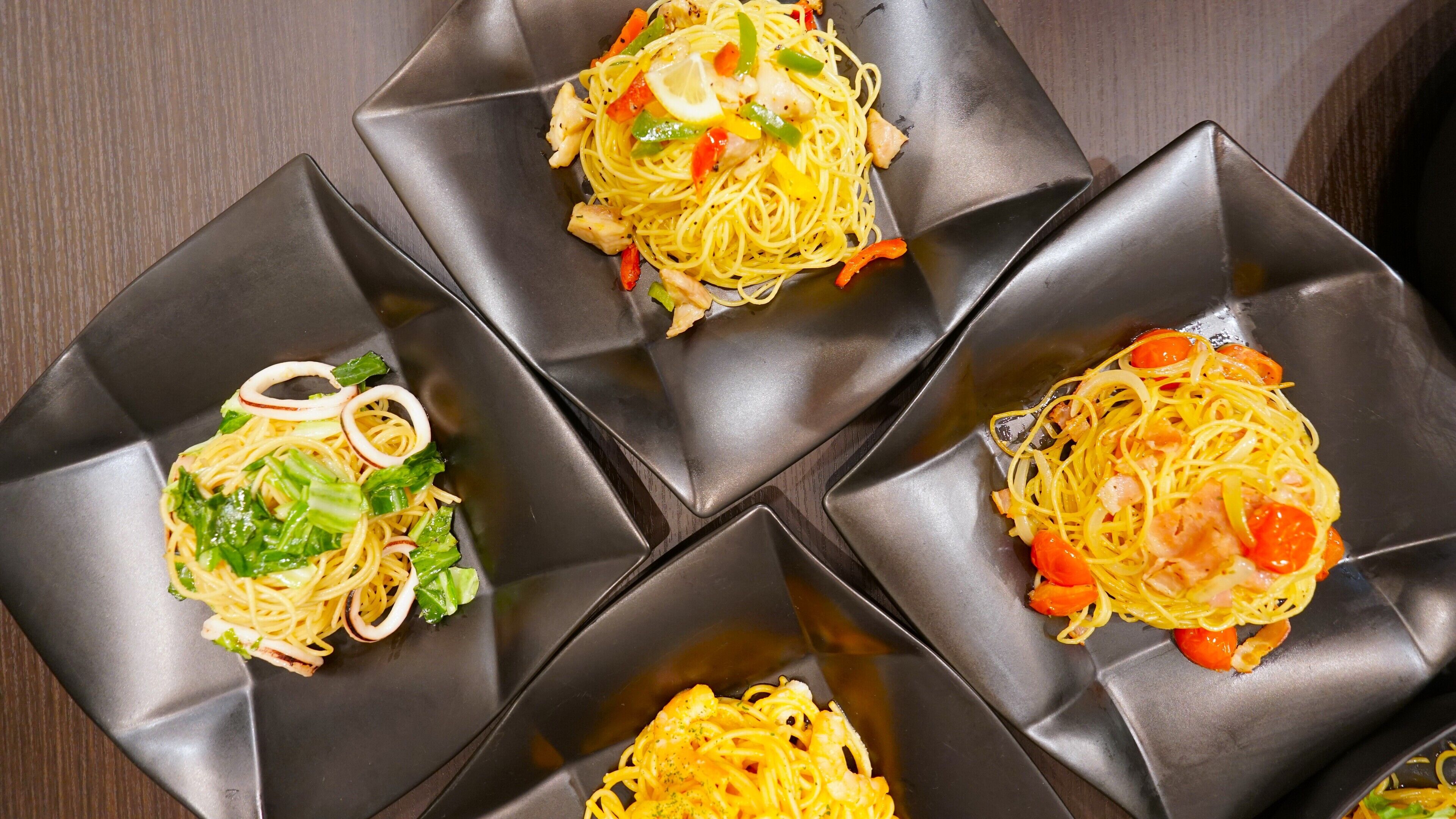 Quatre assiettes carrées de spaghettis aux légumes et au poulet, disposées sur une surface sombre.