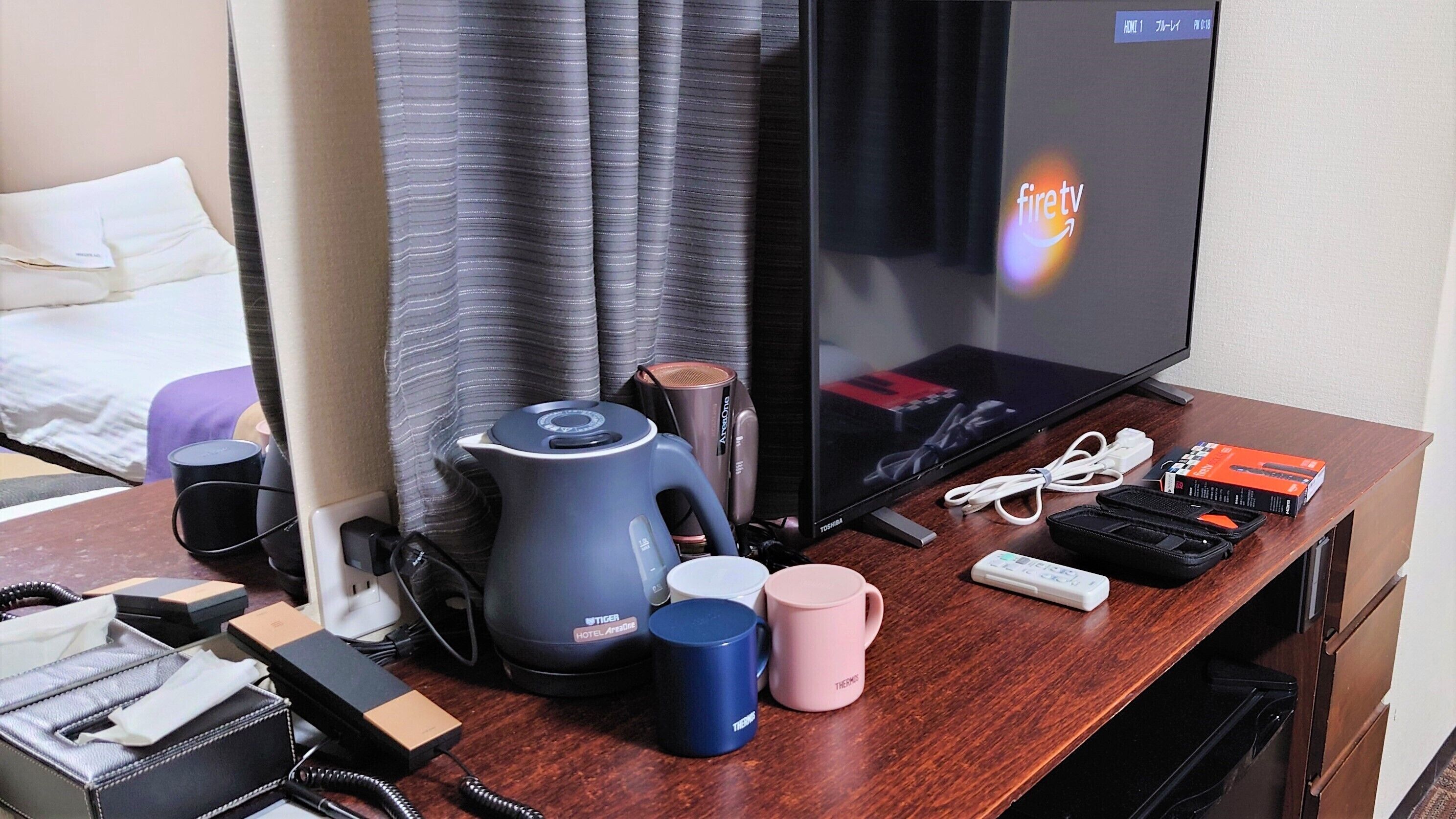 A hotel room with a desk featuring a Toshiba TV, a kettle, a coffee maker, a phone, a remote, and a charging station.