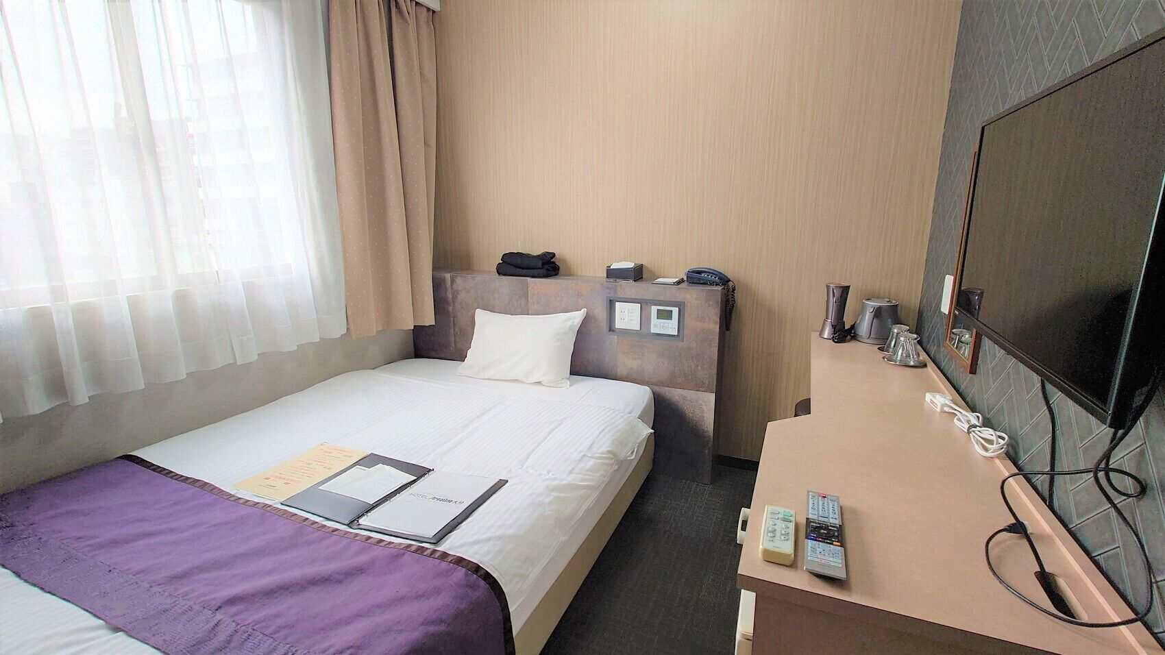 A hotel room with a bed, a desk with a telephone, a television, and a window with curtains.