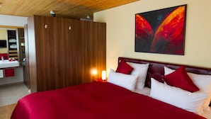 A hotel room with a wooden ceiling, a bed with red bedding, a nightstand with a lamp, and a painting on the wall.