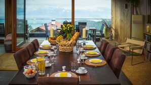 A dining table set for eight with plates, cutlery, and glasses, surrounded by leather chairs, with a view of mountains through large windows.