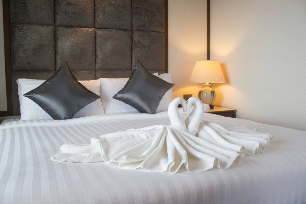 A neatly made bed with two pillows, a lamp on a bedside table, and towels arranged on the bed.