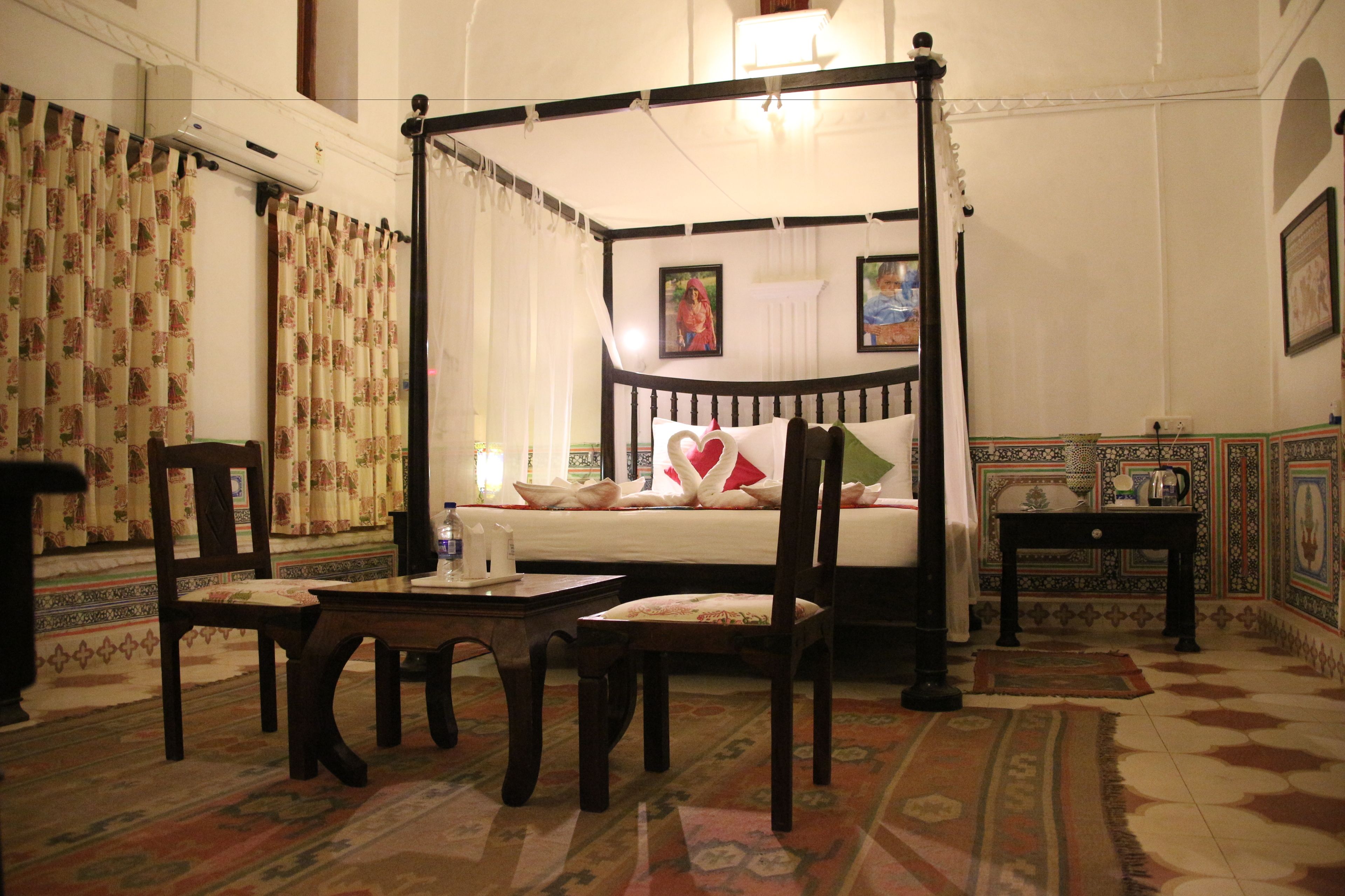 A four-poster bed with a white bedspread and pillows, a wooden nightstand, a table with a chair, patterned curtains, and a decorative wall with a framed picture.