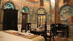 Una habitación con cama, dos sillones, una mesa y decoraciones ornamentales en las paredes, incluyendo murales y motivos florales.