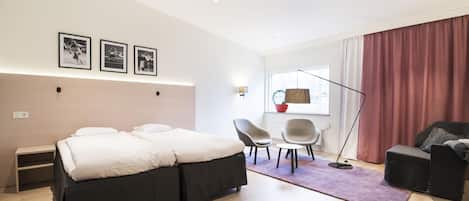 A modern hotel room with a large bed, two chairs, a small table, and a window with curtains.