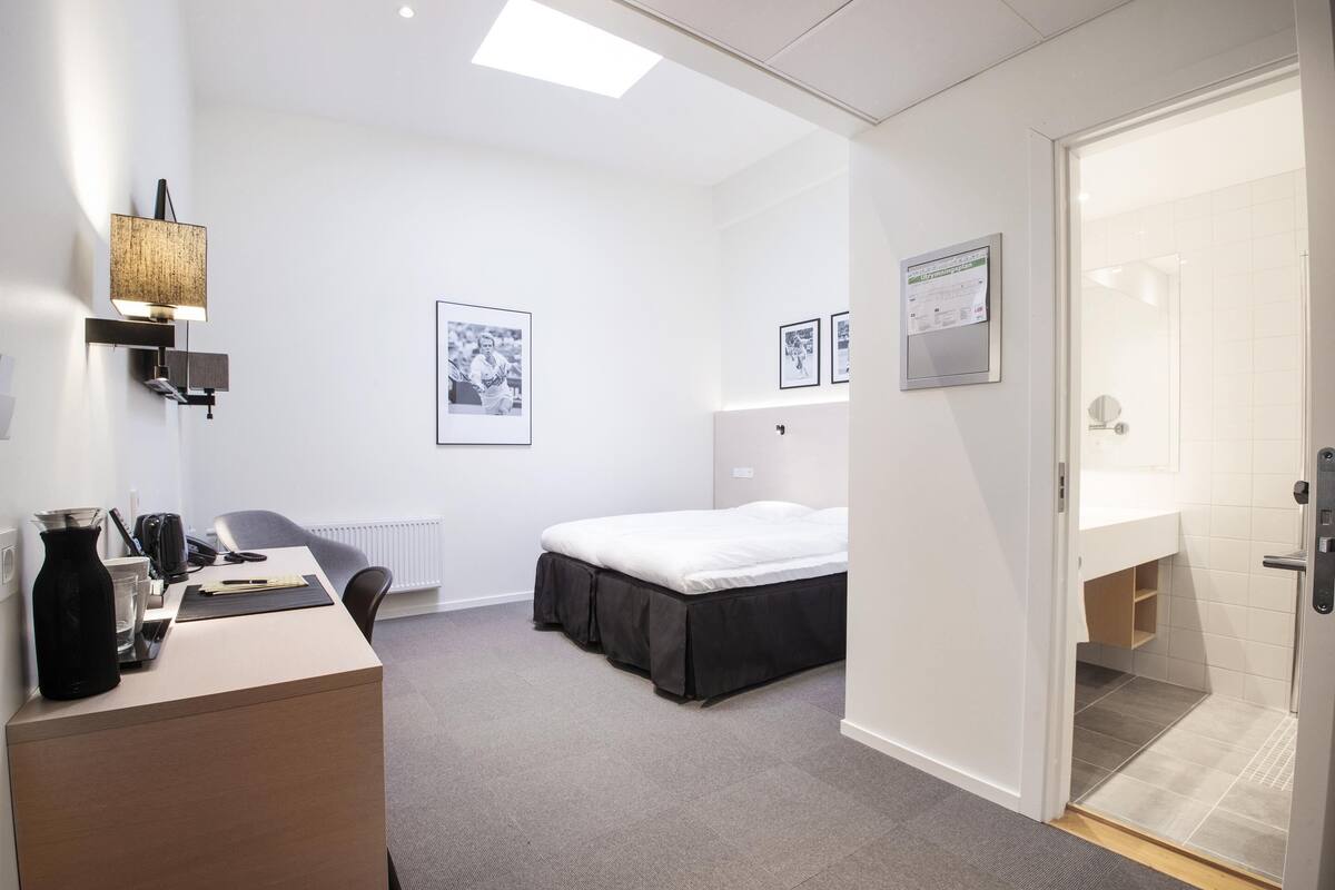 A modern hotel room with a bed, a desk with a lamp, a radiator, and framed pictures on the wall.
