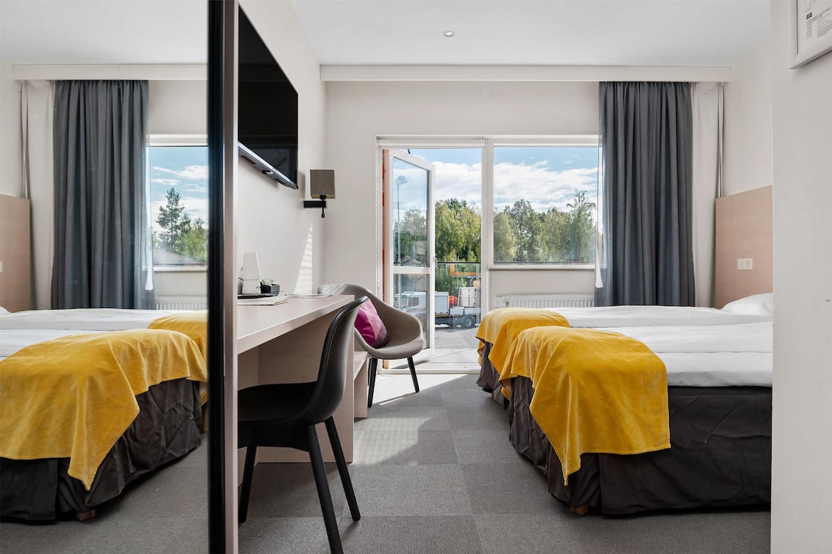 A hotel room with two beds, a desk, and a chair. There is a view of the outdoors through large windows.