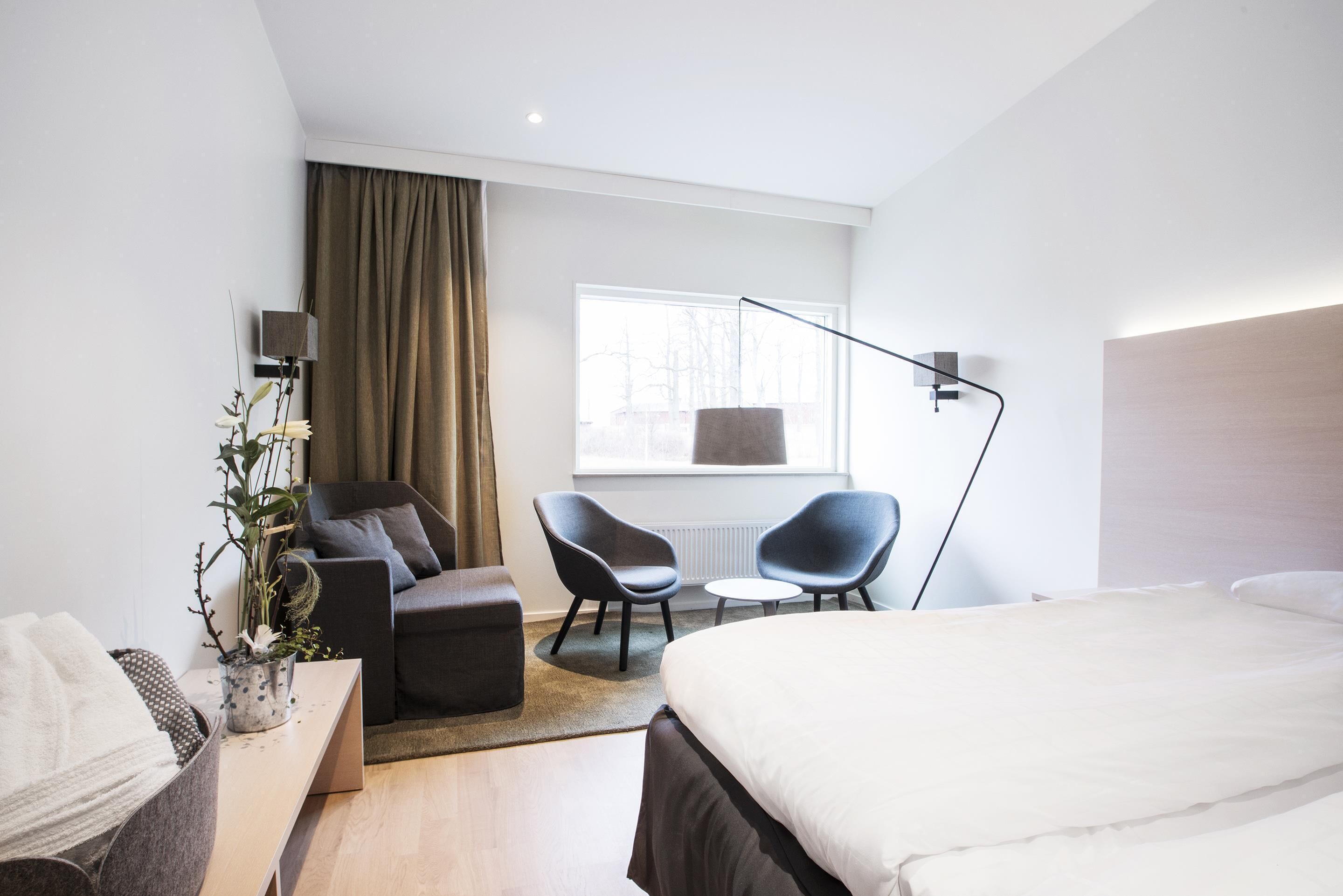 A modern hotel room with a bed, two armchairs, a small table, a potted plant, and a window with curtains.
