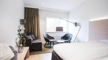A modern hotel room with a bed, two armchairs, a small table, a potted plant, and a window with curtains.