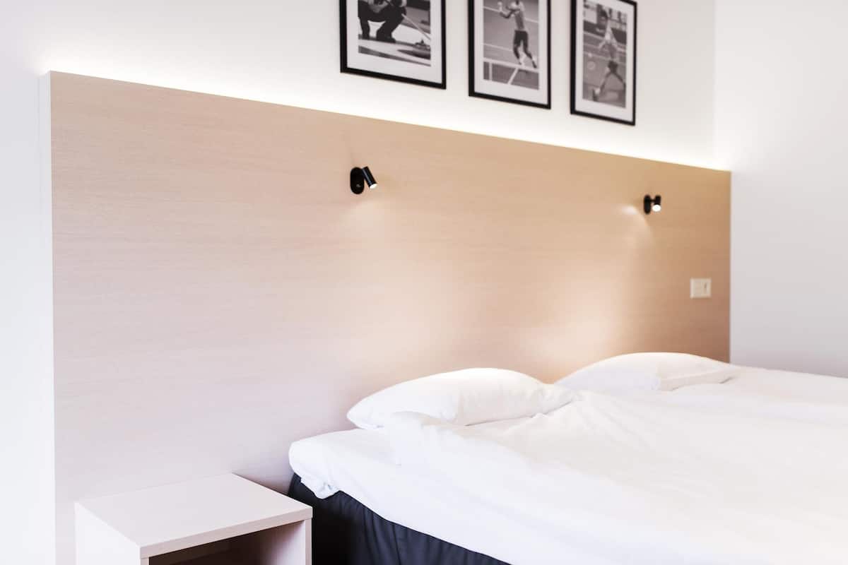 A neatly made bed with white linens, a wooden headboard, and two wall-mounted light fixtures.