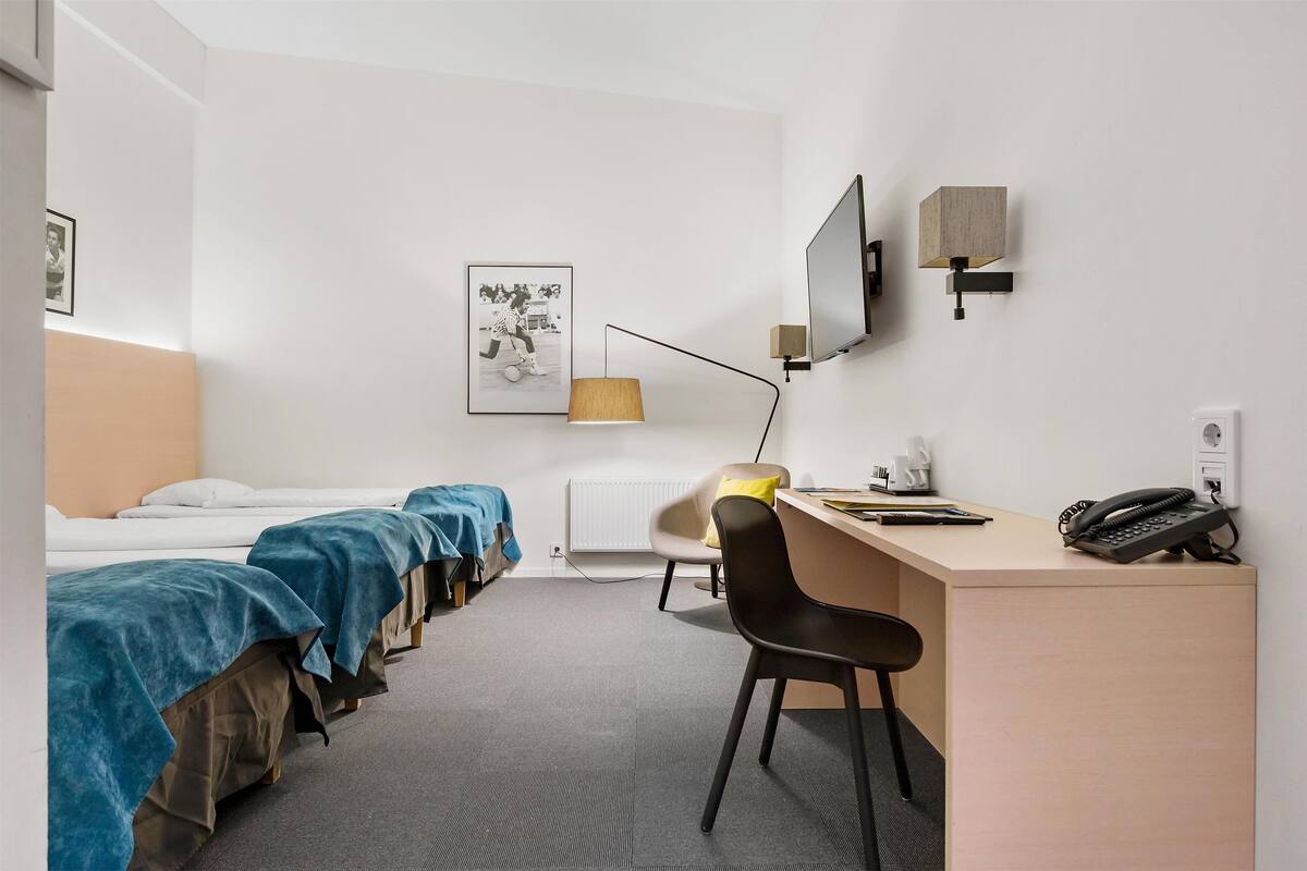A hotel room with two beds, a desk with a chair, a telephone, a lamp, a television, and a radiator.