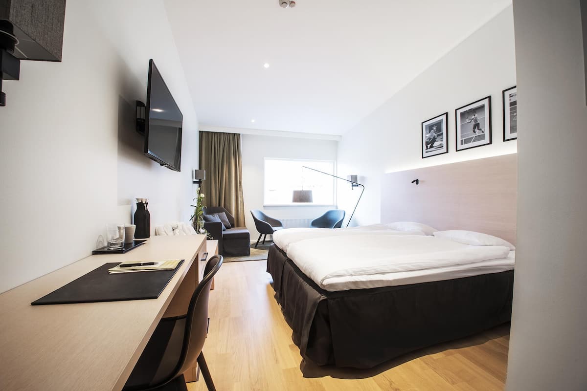 A modern hotel room with a bed, desk, chair, TV, and artwork on the wall.