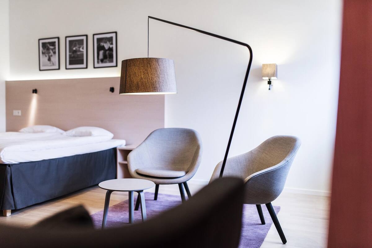 A modern hotel room with a bed, two chairs, a small table, a floor lamp, and wall-mounted pictures.