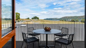 Un balcon doté d’une table ronde et de quatre chaises, offrant une vue panoramique sur un paysage pittoresque avec des collines et une étendue d’eau.