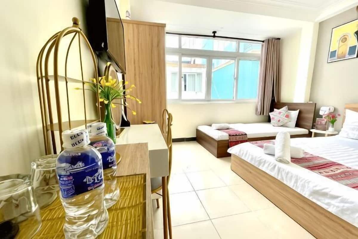 Ein Hotelzimmer mit einem Bett, einem Schreibtisch mit Stuhl, einem Fenster mit Vorhängen, einem kleinen Tisch mit Wasserflaschen und einer Vase mit Blumen.