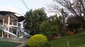 A modern two-story house with a curved roof, a staircase leading to the upper floor, and a well-maintained garden with various plants and a small bush. - Motsamai Lodge (Bulawayo)