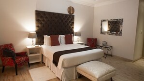 A bedroom with a tufted headboard, a large bed with multiple pillows, two bedside tables with lamps, a red chair, a small table with a mirror, and a bench. - Motsamai Lodge (Bulawayo)