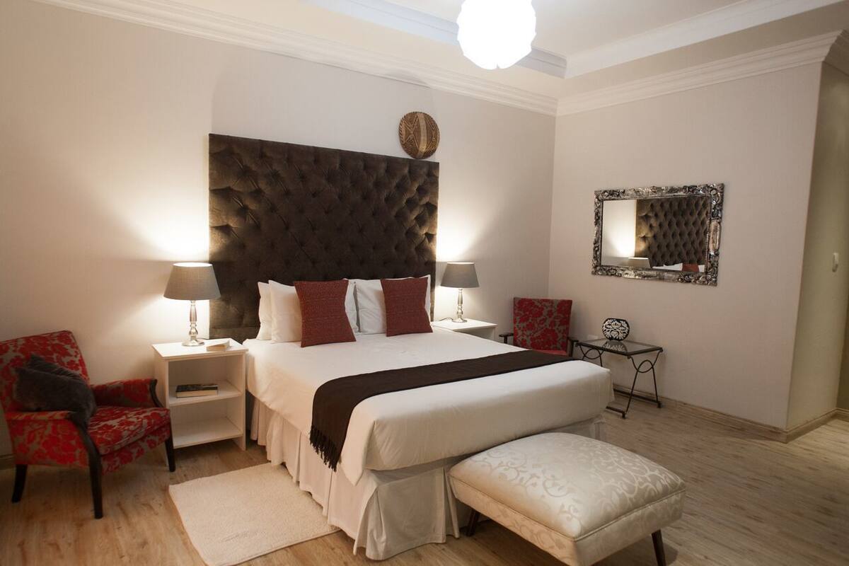 Un dormitorio con un cabecero capitonado, una cama grande con muchos almohadones, dos mesitas de noche con lámparas, una silla roja, una pequeña mesa con espejo y un banco.