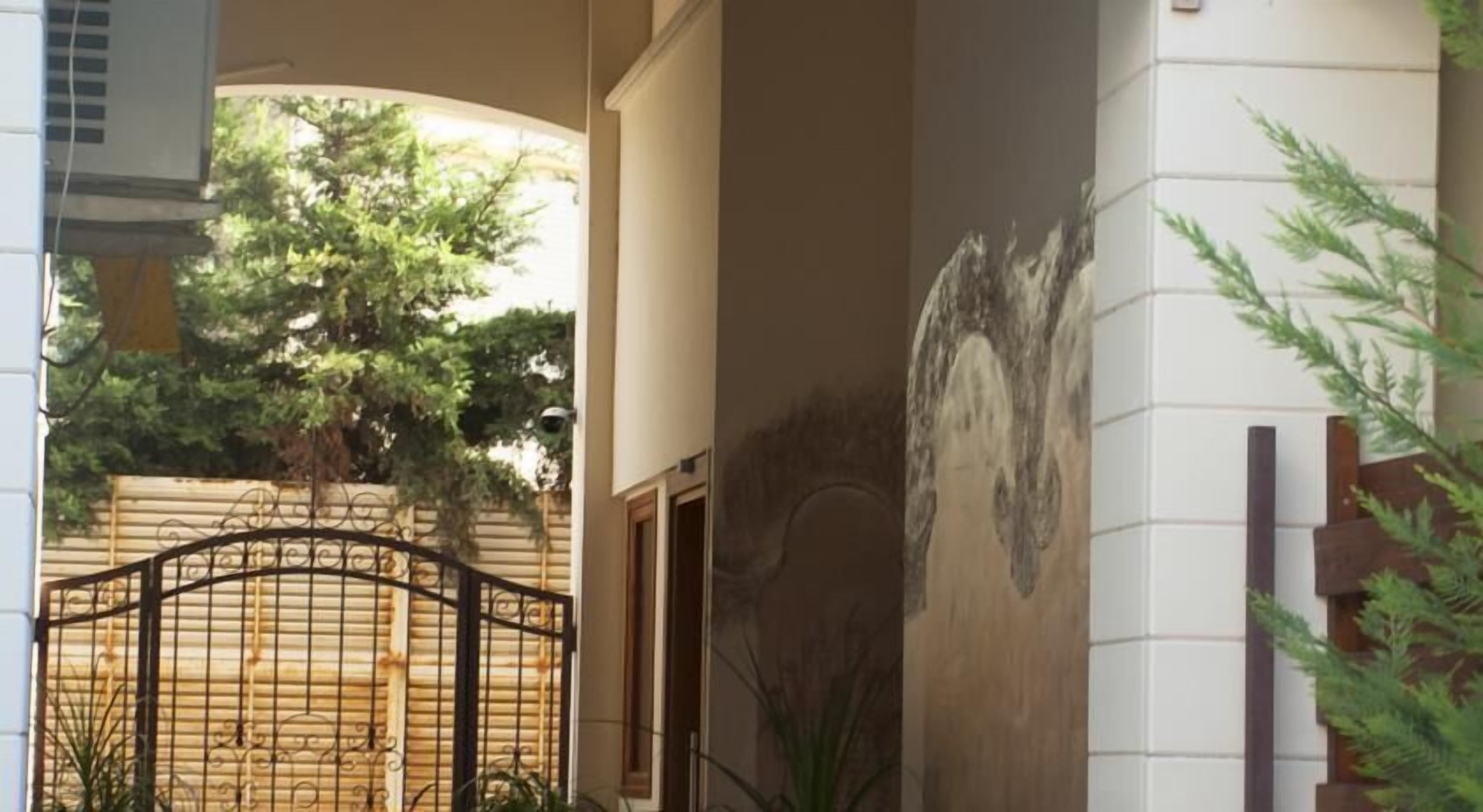 Una entrada de edificio con una reja metálica, un muro con un mural y un árbol al fondo.