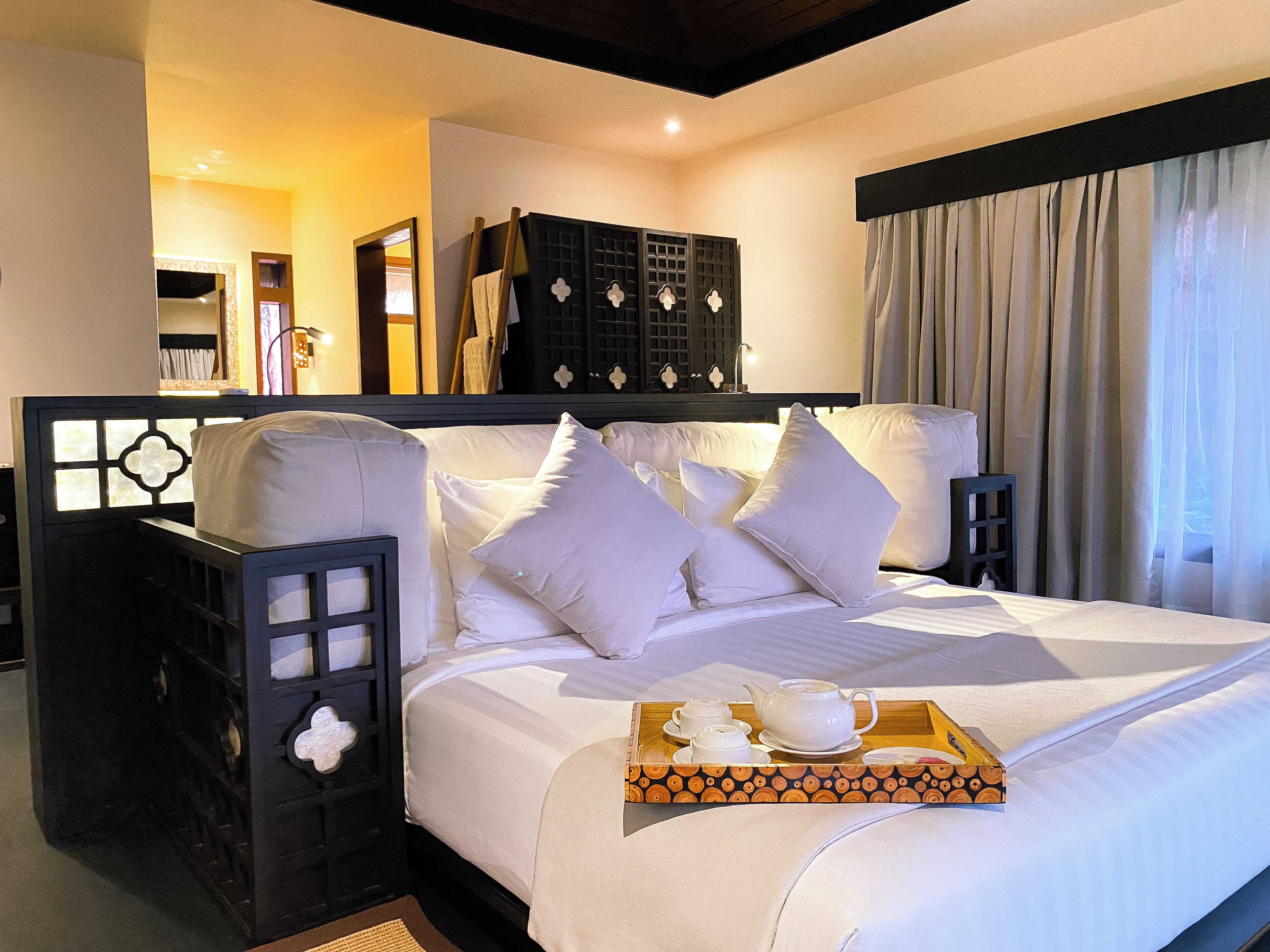 A neatly made bed with white linens and pillows, a tray with tea set, and a dark headboard with a geometric pattern.