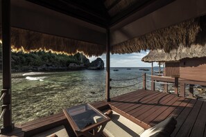 Una terraza de madera con una mesa de vidrio, techo de paja y vista al mar y la costa rocosa.