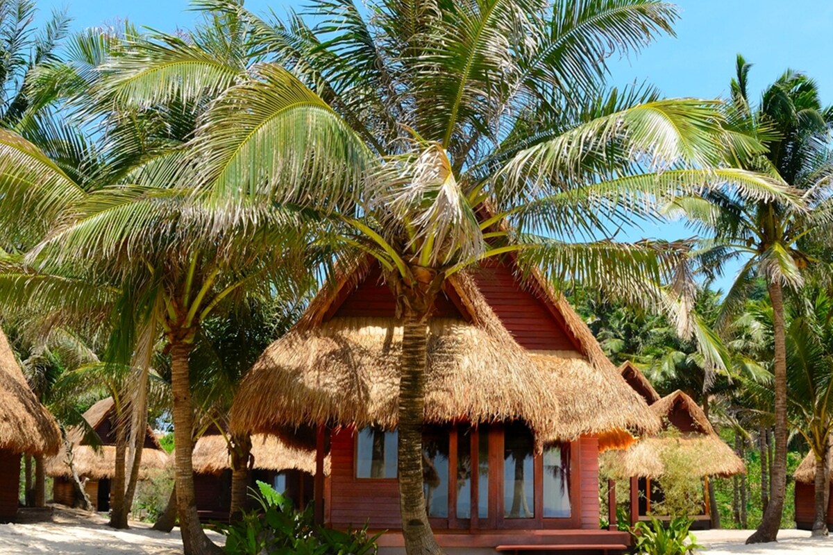 A thatched-roof bungalow with a red exterior, surrounded by palm trees and other tropical vegetation.
