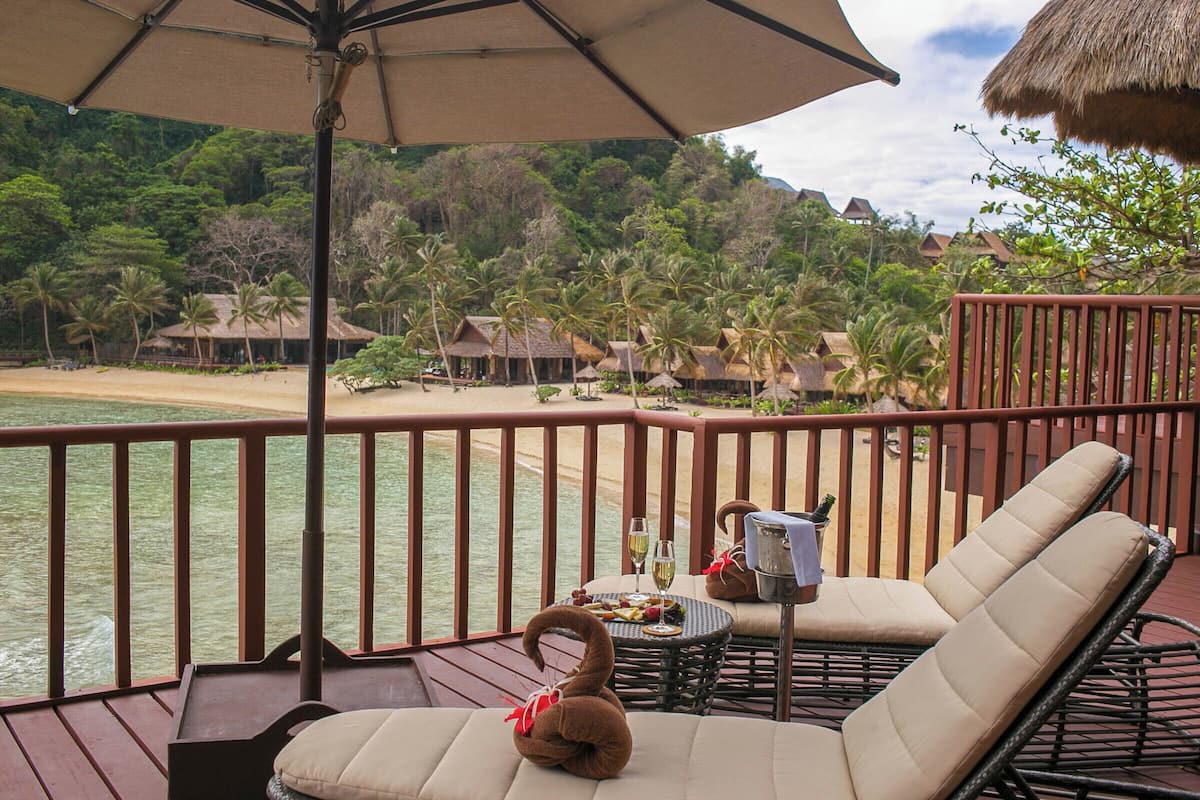 Eine Terrasse mit Liegestühlen, einem Tisch mit Flasche und Gläsern und Blick auf einen Strand sowie Gebäude mit Strohdächern.