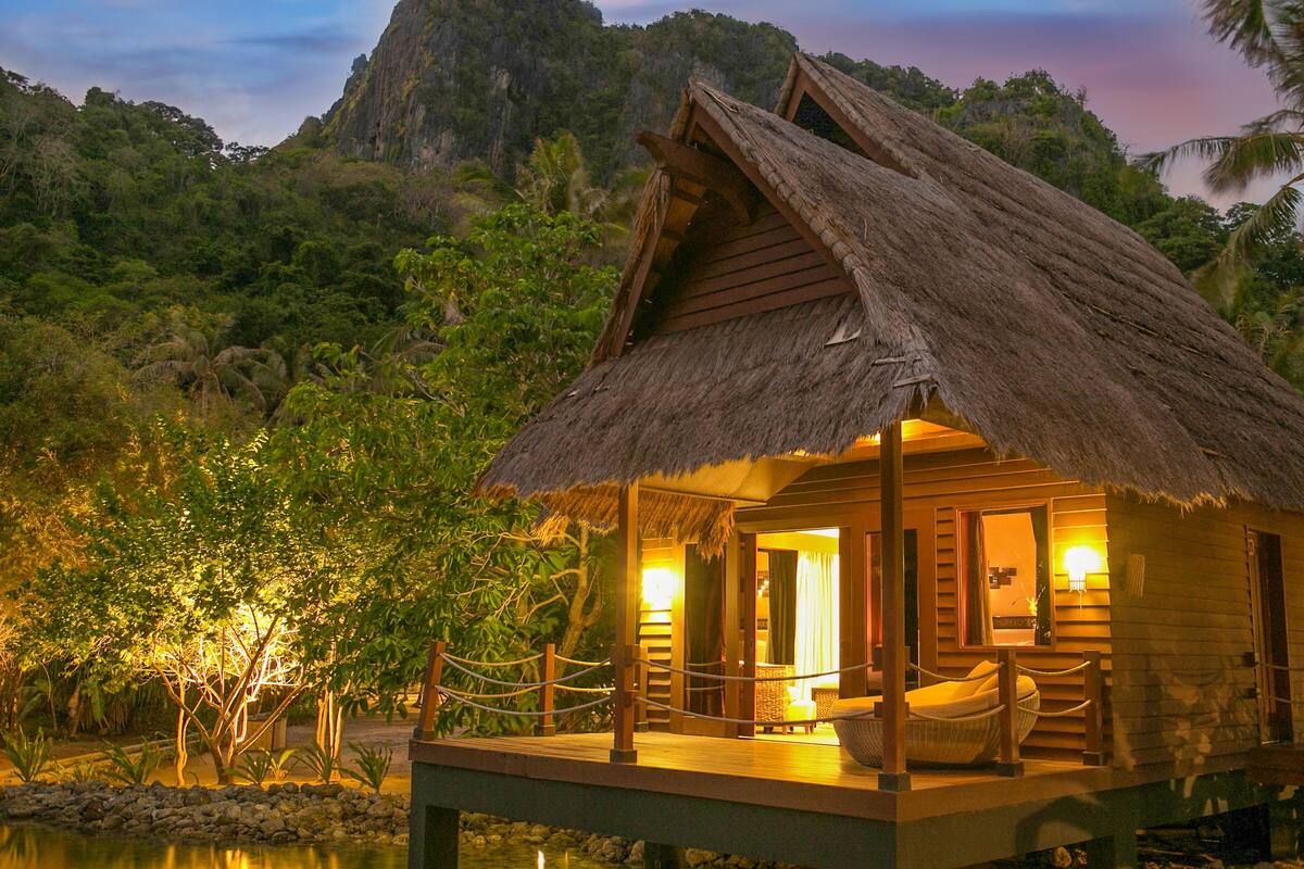 Ein reetgedecktes Bungalow mit einer Holzdauerwiesenfläche, einer Hängematte und Blick auf den umliegenden Wald und das Wasser.