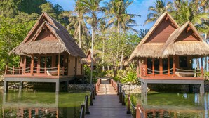 A wooden bridge connecting two thatched-roof overwater bungalows with a clear view of palm trees and a calm water body. - Cauayan Boutique Private Island (El Nido)