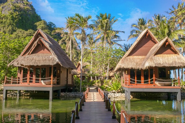 A wooden bridge connecting two thatched-roof overwater bungalows with a clear view of palm trees and a calm water body. - Cauayan Boutique Private Island (El Nido)