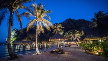 A beach with palm trees, a thatched-roof hut, and a lounge chair.